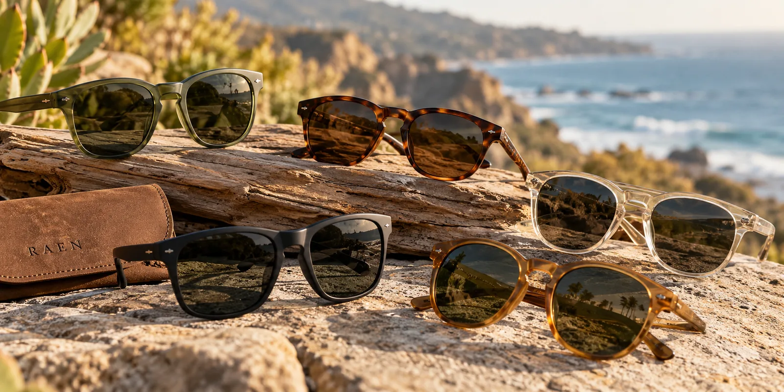 Several RAEN sunglasses frames are arranged on a coastal rock beside a branded case.
